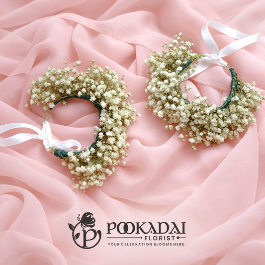 Pair of ivory-white baby’s breath hand gajras with white ribbon ties on blush pink fabric, with Pookadai Florist branding.